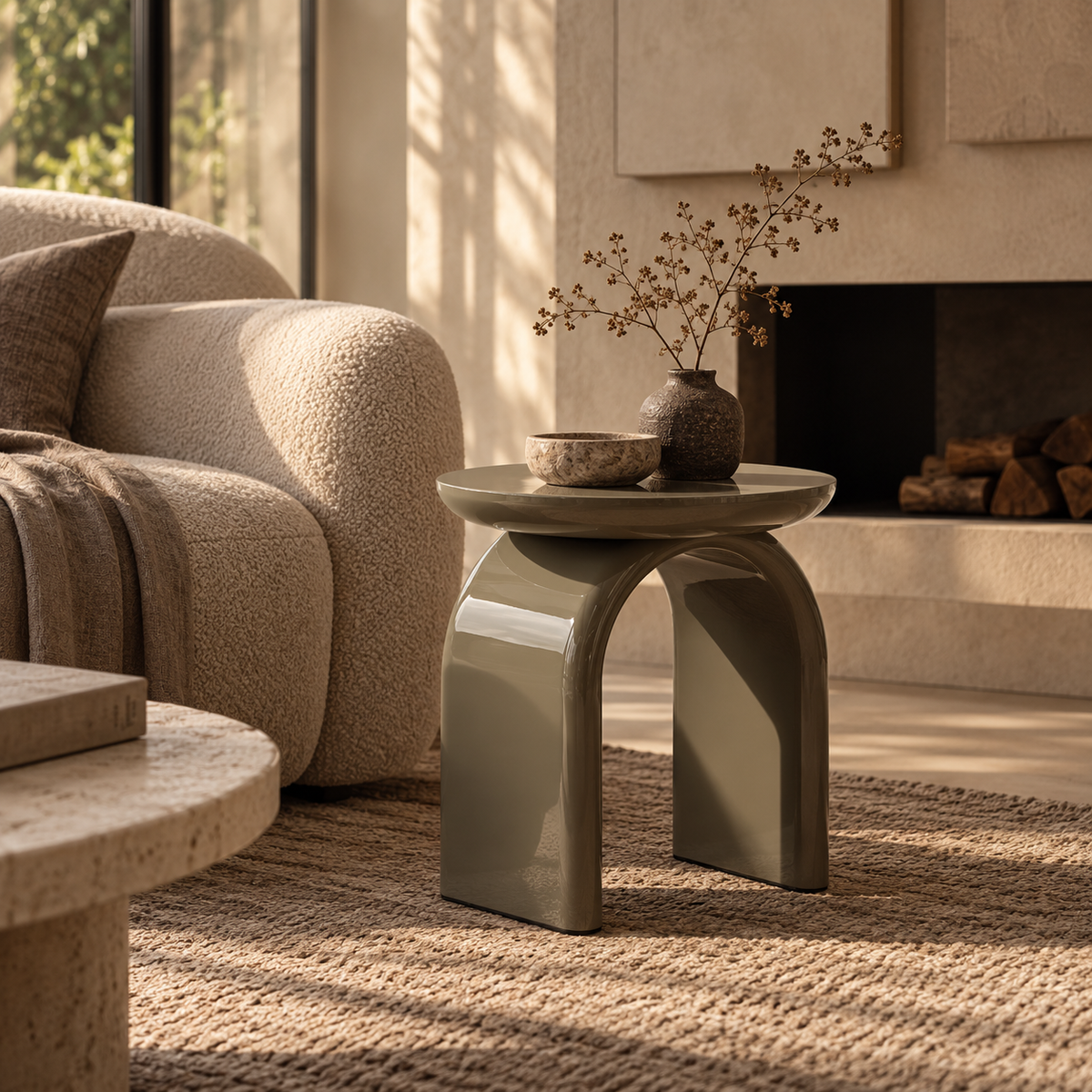 Beistelltisch Alois in glänzendem Greige mit skulpturaler Bogenform, inszeniert in einem warmen, hochwertigen Wohninterieur mit natürlichen Materialien und dezenter Dekoration